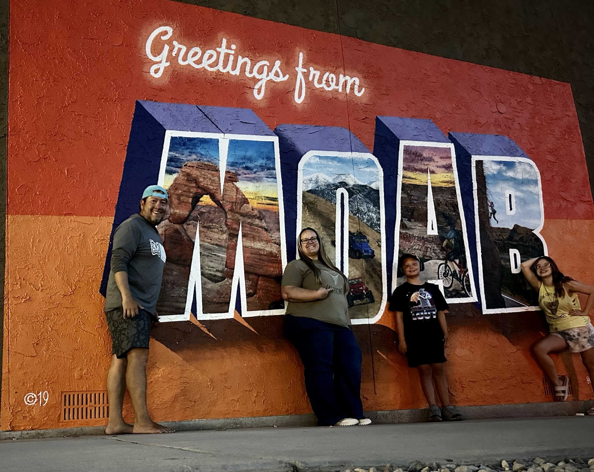 A family stands in front of a mural that says Greetings from Moab